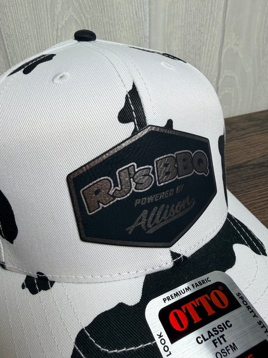 A white and black cow-print snapback cap featuring a black patch on the front that reads 'RJ's BBQ Powered by Allison'. A white retail sticker for 'OTTO' is visible on the brim.