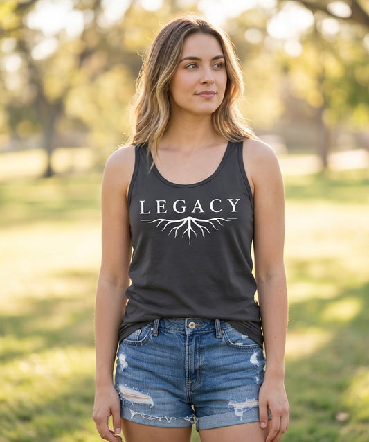 Woman wearing a black tank top with 'LEGACY' printed on it, standing in a park.