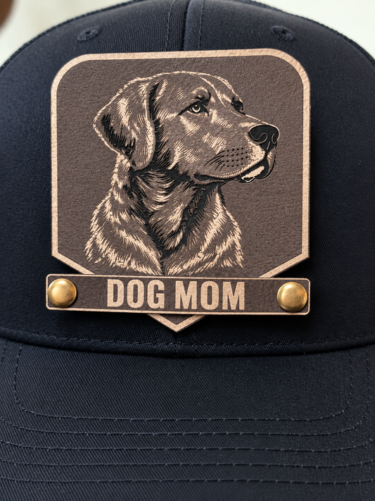 A close-up of a dark navy baseball cap featuring a large embroidered patch with a black Labrador retriever illustration and the words 'DOG MOM' in white text on a tan background, secured with two gold-tone snap buttons.
