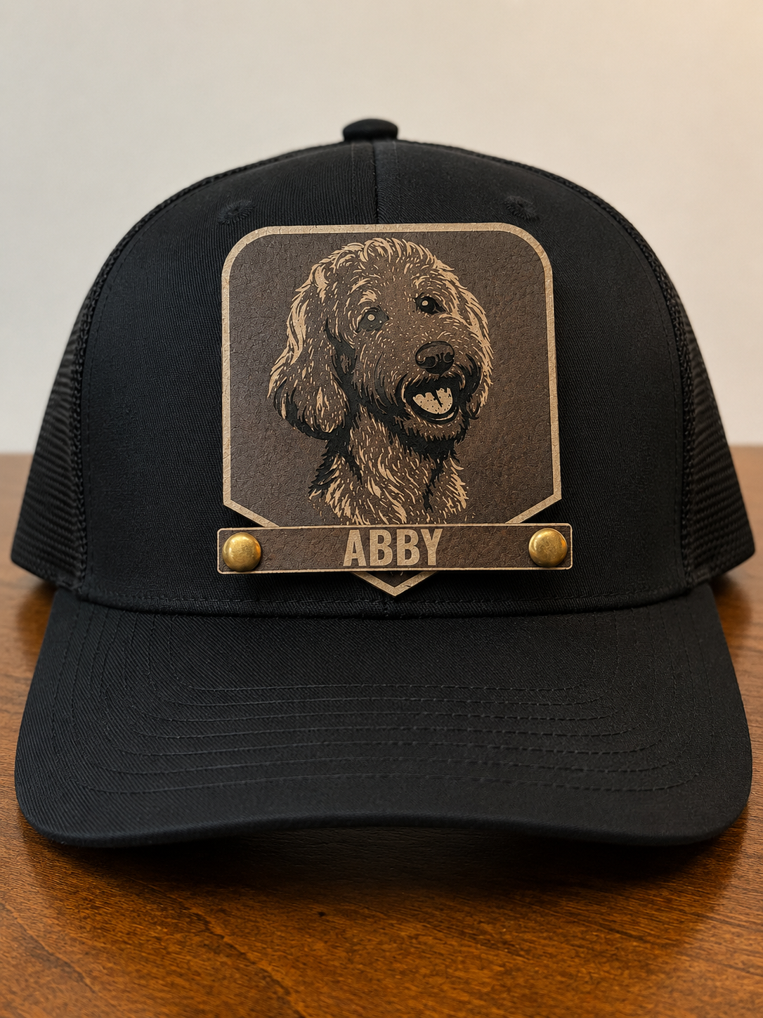 A black trucker-style cap with a custom rectangular patch featuring a dog portrait and the name 'ABBY' in gold-tone lettering, displayed on a wooden surface against a neutral background.