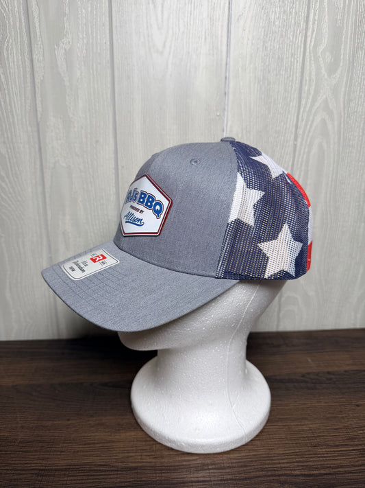 A gray trucker-style baseball cap with a blue, white, and red American flag pattern on the mesh back, displayed on a white mannequin head against a light wood-paneled wall.