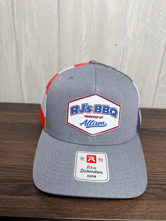 A heather gray trucker hat with red and navy mesh side panels, featuring a 'RJ's BBQ Powered by Allison' patch on the front and a Richardson 05FM sticker on the brim, displayed on a wooden surface against a light wood paneled wall.