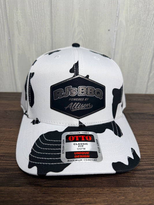 A white and black camouflage snapback cap with a black hexagonal patch reading 'RJBBQ Powered by Allison' on the front, resting on a wooden surface against a light wood paneled background.