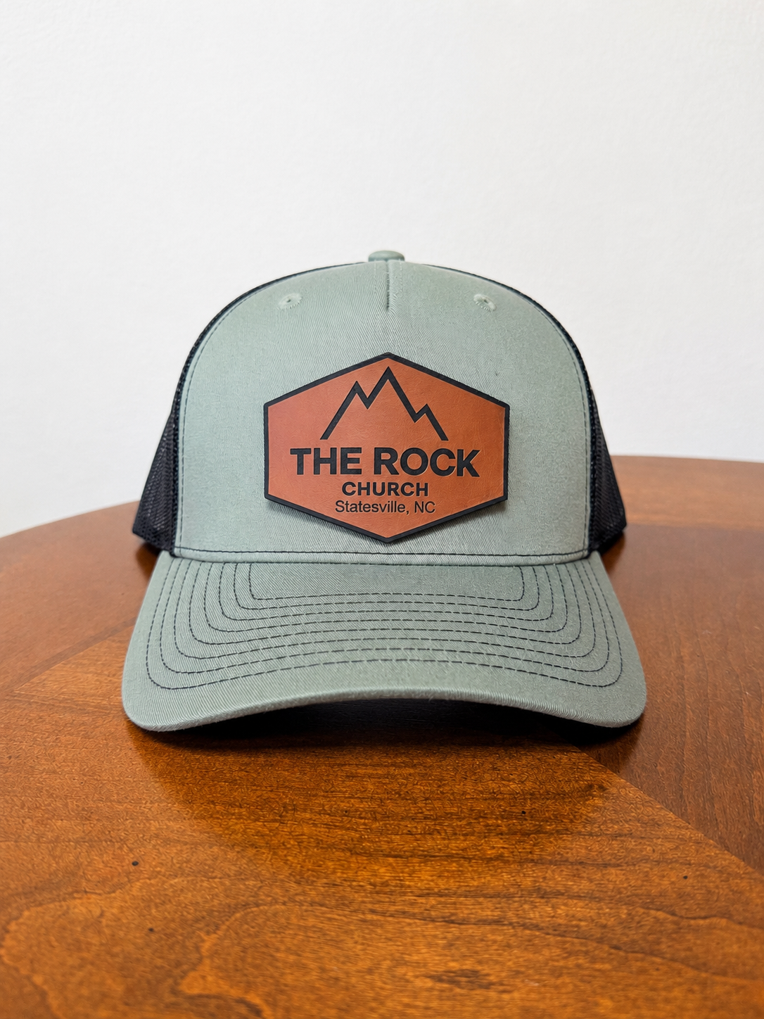 A sage green and black trucker hat with a brown and black 'The Rock Church' patch on the front, resting on a wooden table against a white wall.
