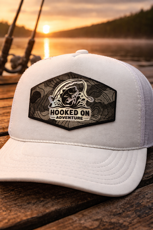 White cap with 'Hooked on Adventure' patch on a wooden surface with a sunset over water background
