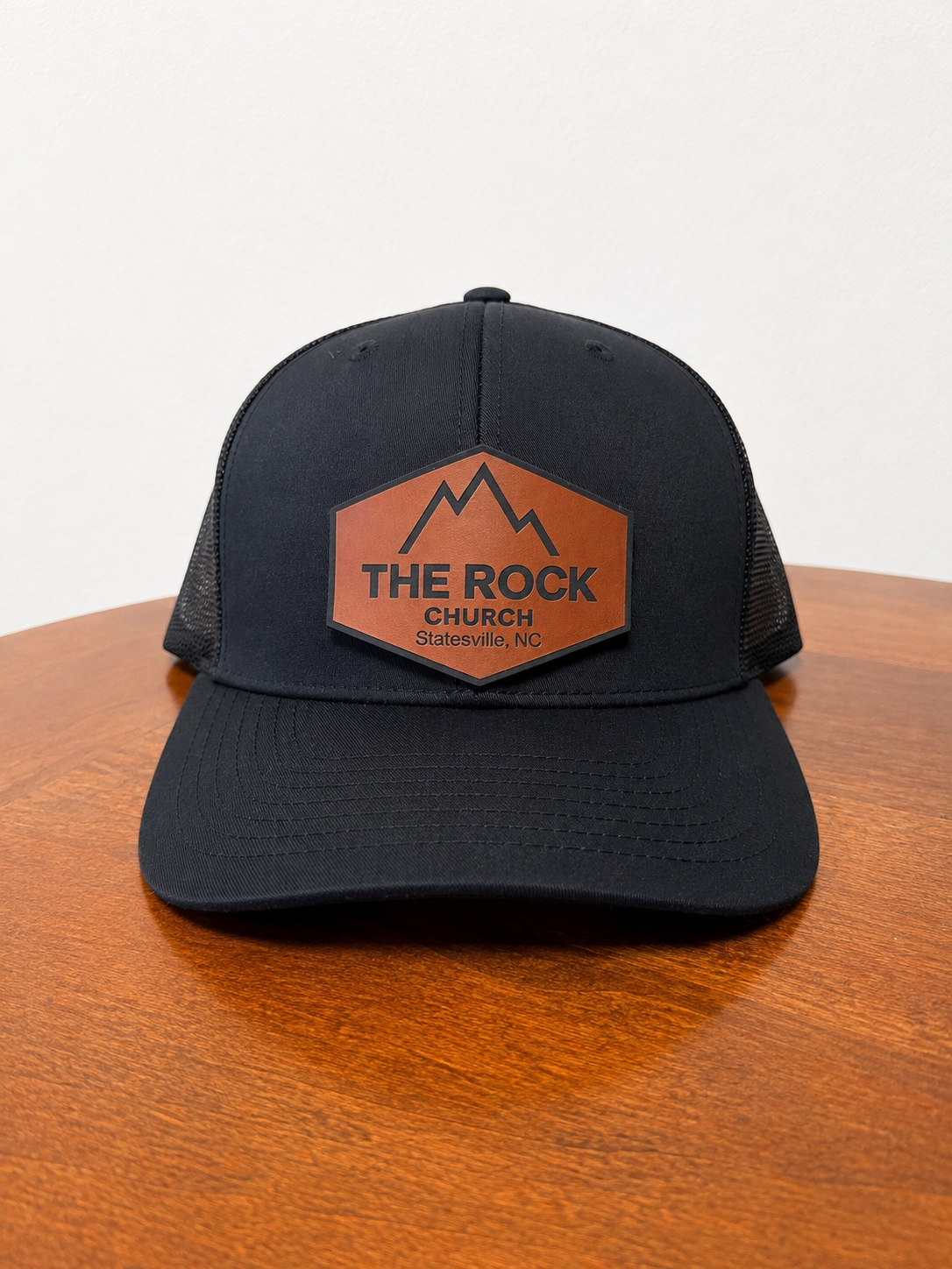 A black trucker-style cap with a brown leather patch on the front that reads 'THE ROCK CHURCH' and 'Saratoga, NC' beneath a mountain graphic, displayed on a wooden table against a light background.