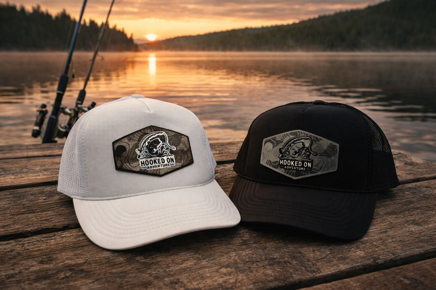 Two hats with 'Hooked On' logo on a wooden dock by a lake at sunset.