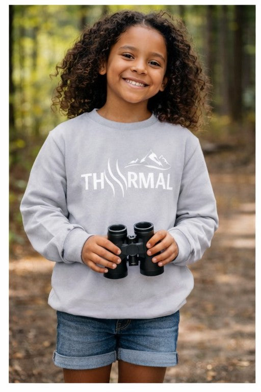 Child wearing a gray sweatshirt with 'THRMAL' logo, holding binoculars in a forest setting
