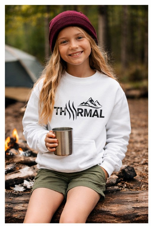 Child wearing a white sweatshirt with THRMAL logo, holding a mug by a campfire in a forest.