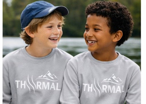 Two children wearing gray sweatshirts with a logo sitting on a rock by a body of water.