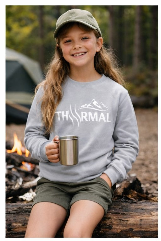 Child wearing a sweatshirt with 'The Rival' logo, holding a mug, sitting by a campfire in a forest.
