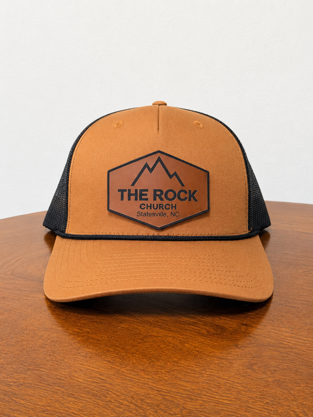 A tan and black trucker hat with a brown leather patch on the front that reads 'THE ROCK CHURCH Statesville, NC', displayed on a wooden table against a white wall.